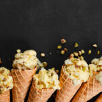 El Buen Aroma Cafetería Heladería - Cafetería en Andújar Vanilla ice cream in waffle cone with nuts on black stone background.
